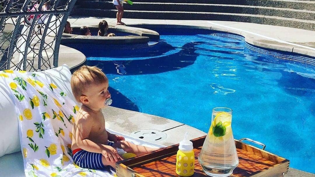 A baby sitting by the poolside with a lemon hooded towel at the back